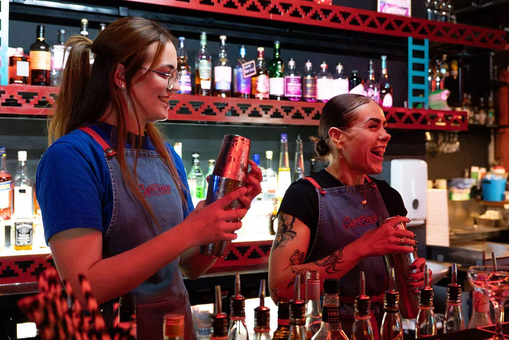 Two Bartronica bartenders laughing while shaking cocktails behind the bar