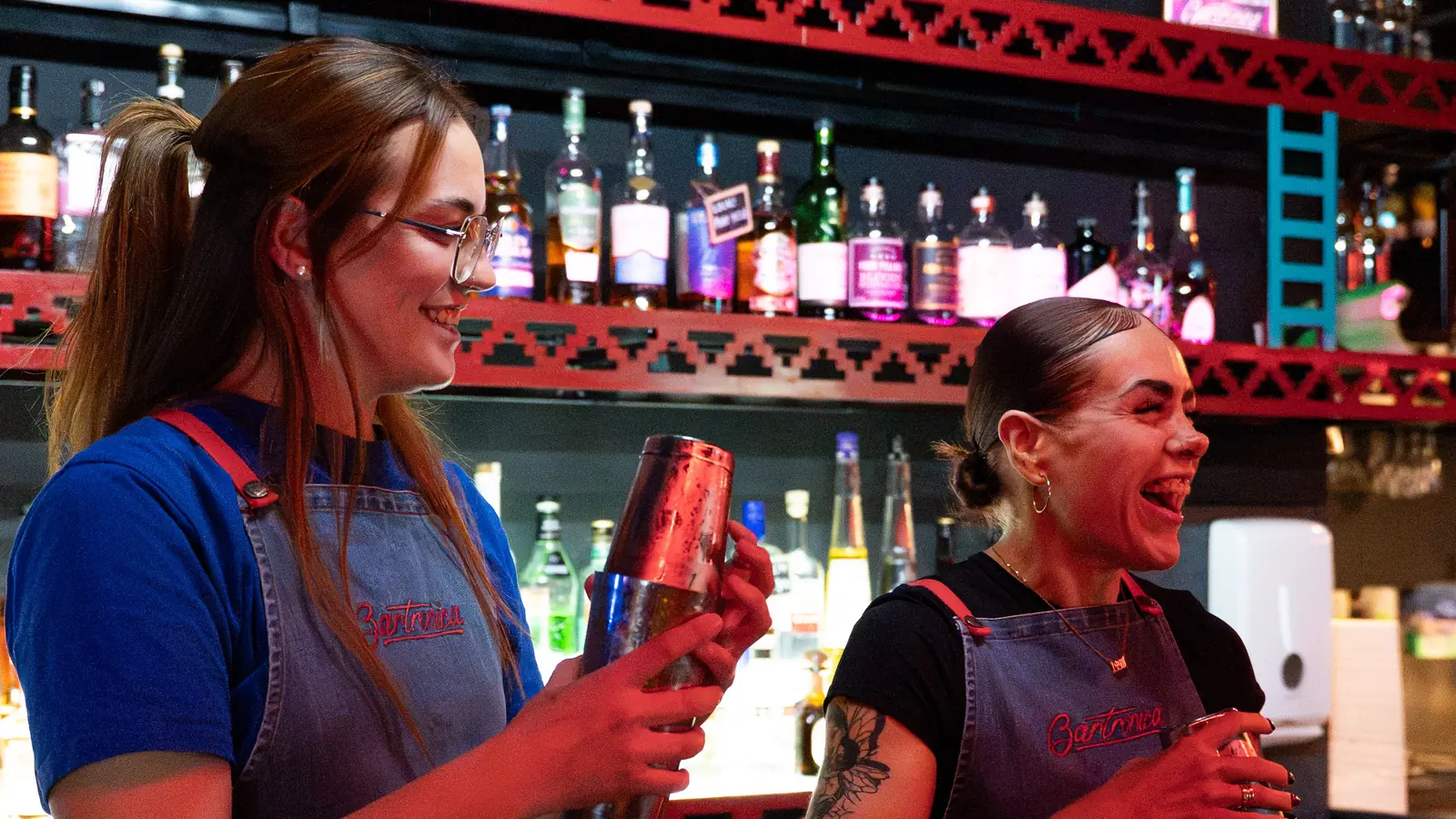 Two Bartronica bartenders laughing as they shake cocktails in front of the neon-lit back bar